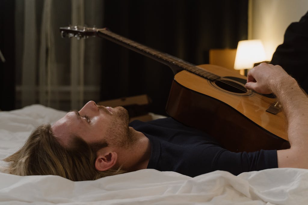 A man lying in bed with an acoustic guitar, reflecting contemplation after getting support at musician therapy collective