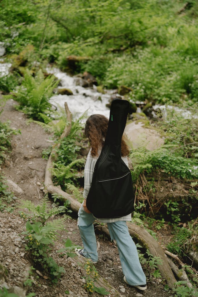 A woman carrying a guitar is hiking on a lush forest trail beside a stream.
