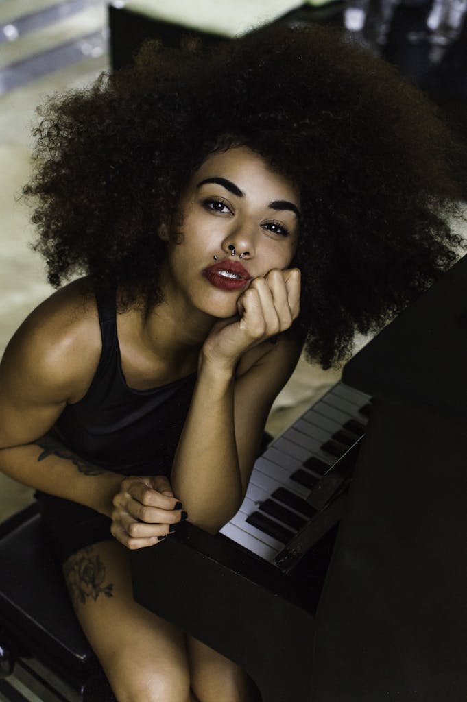 A beautiful woman with curly hair poses at a piano, featuring tattoo and bold red lips.