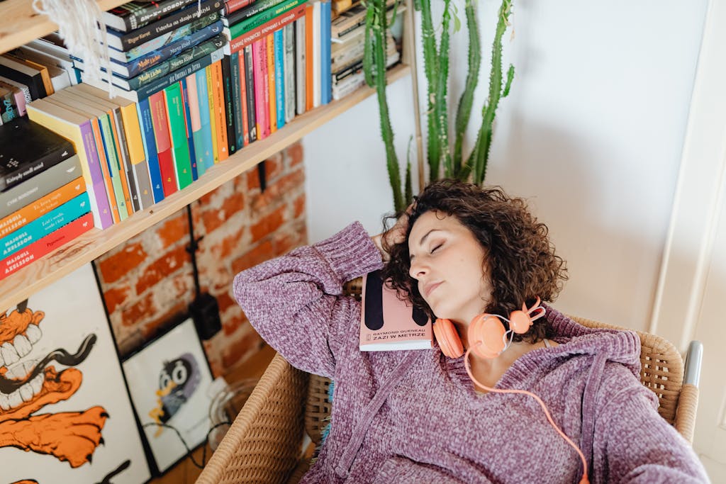 A Caucasian woman with curly hair relaxes at home in a cozy chair, holding a book with headphones.