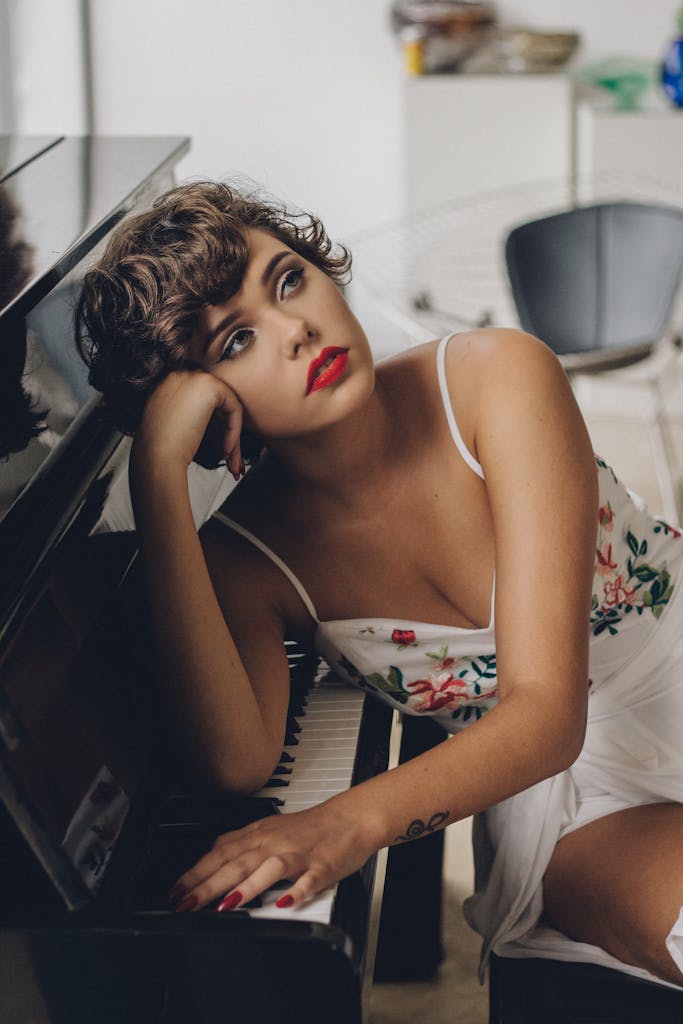 Chic woman in floral dress with red lips posing thoughtfully as she leans on a piano indoors.