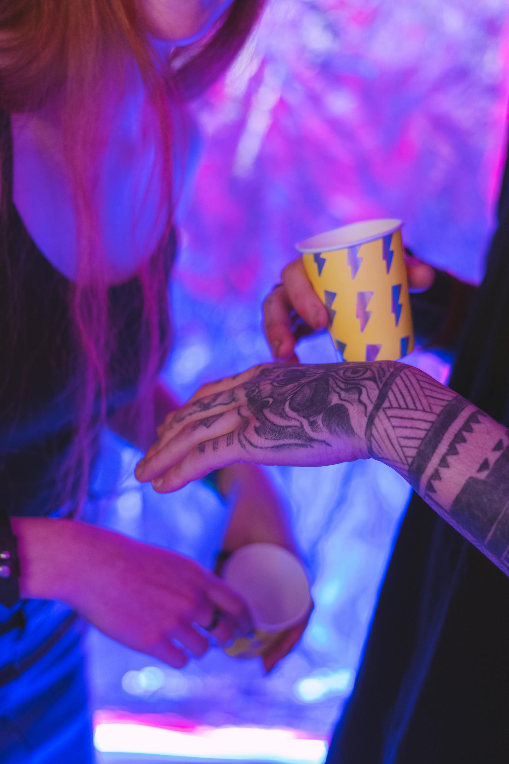 Close-up of tattooed hands holding colorful cups with neon lights in background.