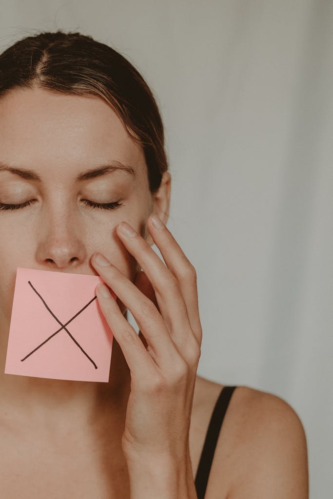 Close-up of woman covering mouth with a note showing an 'X', symbolizing silence or prohibition.