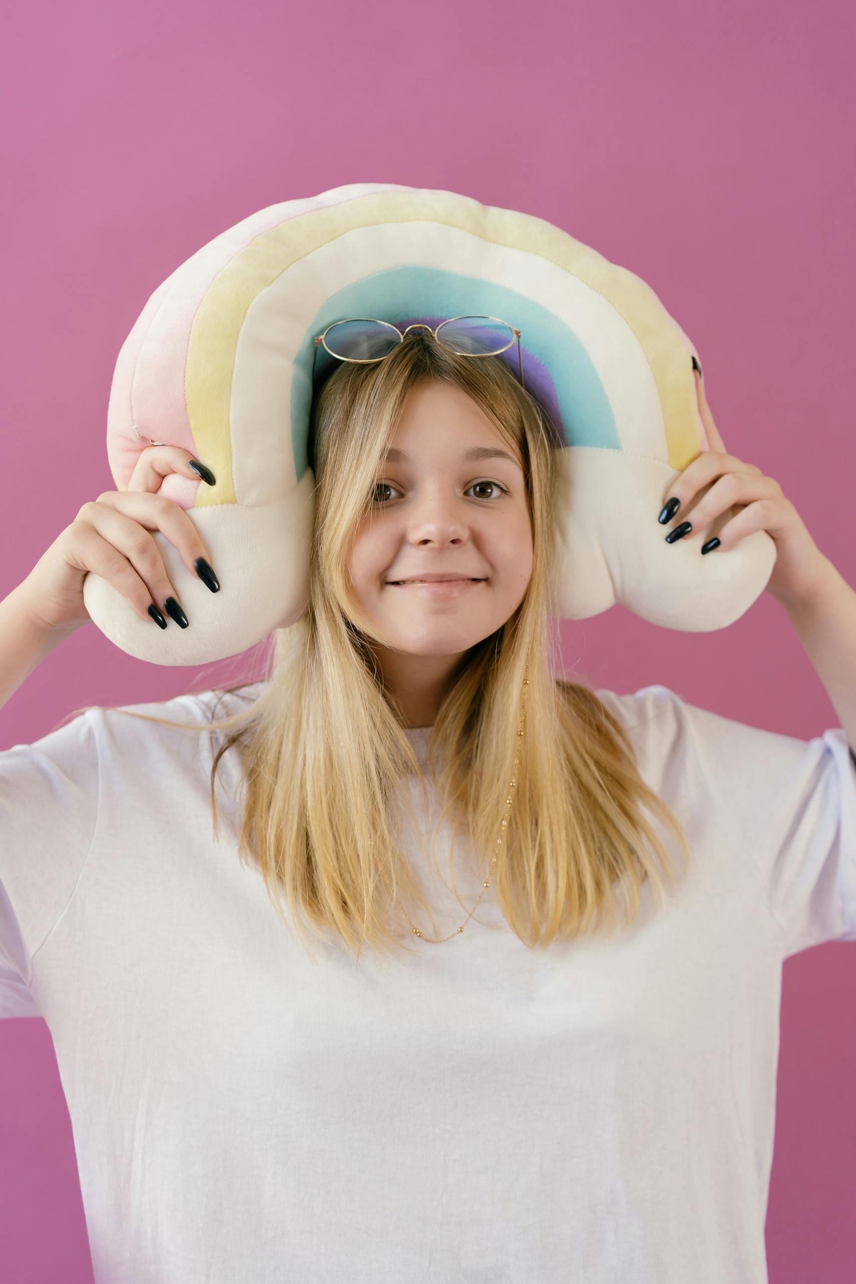 Happy blonde teen posing with a rainbow plush toy against a pink background.