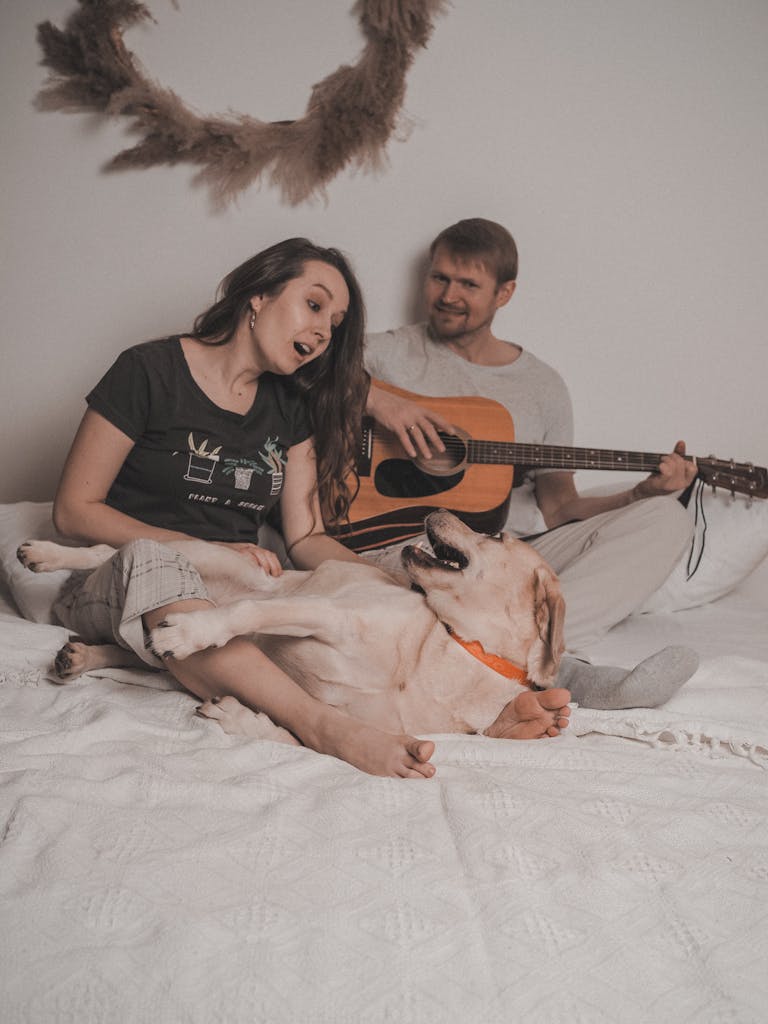 Happy couple playing guitar and singing with their Labrador on bed.
