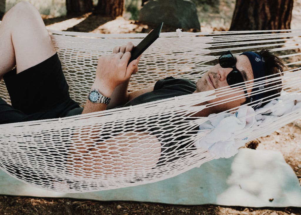 Man relaxing in a hammock outdoors, enjoying a sunny day with sunglasses and a book.