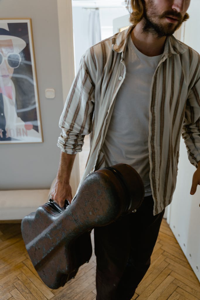 Man with facial hair walking indoors carrying a guitar case in a casual attire.