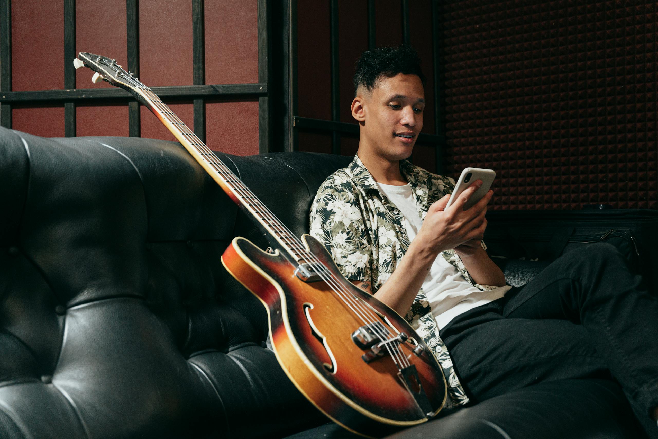 Young man enjoying music break with smartphone and guitar on sofa.