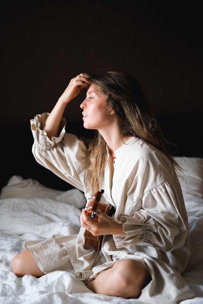 Young woman in a white dress sitting on a bed with a ukulele, enjoying a peaceful moment.